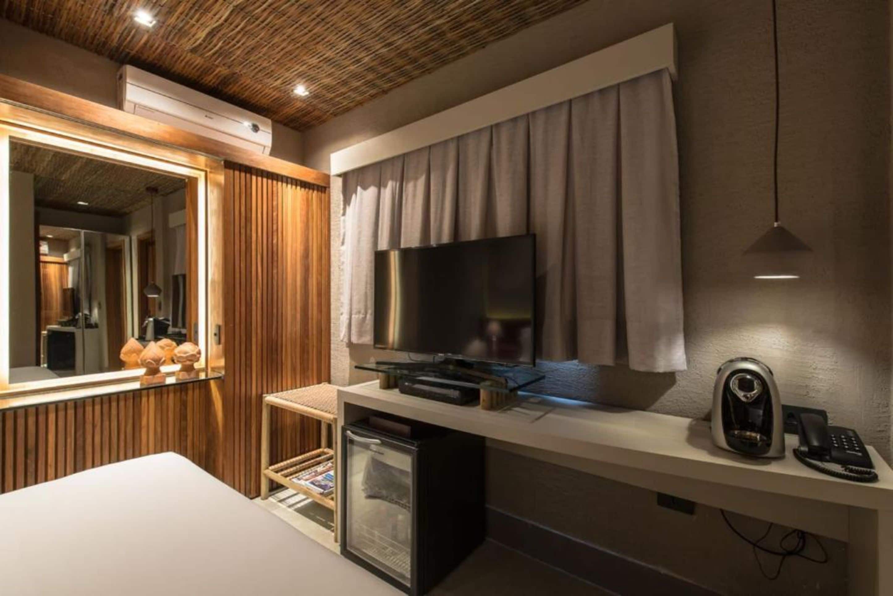 A modern hotel room with a large flat-screen TV, a minibar, a desk with a telephone, and a mirror.