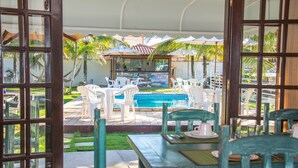 Un área de piscina al aire libre con sillas y mesas de plástico blanco, rodeada de palmeras y sombrillas.