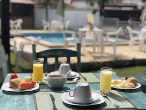 A breakfast table with plates of food, a jug, a cup, and a glass of juice, set outdoors with a pool in the background. - Pousada Casuarina Geribá (Búzios)