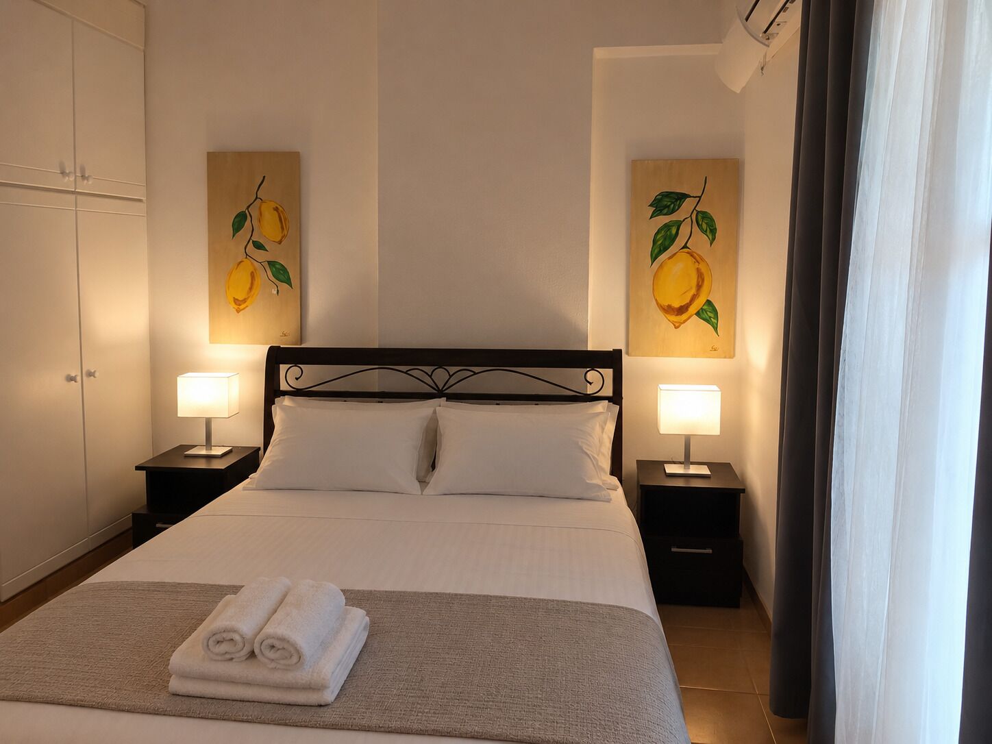 A hotel room with a bed, two bedside lamps, a folded towel on the bed, and two wall-mounted paintings of fruit.