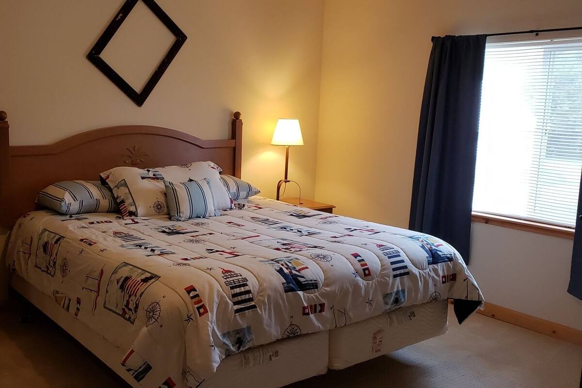 A neatly made bed with a patterned bedspread, a bedside lamp, a nightstand, and a window with curtains.