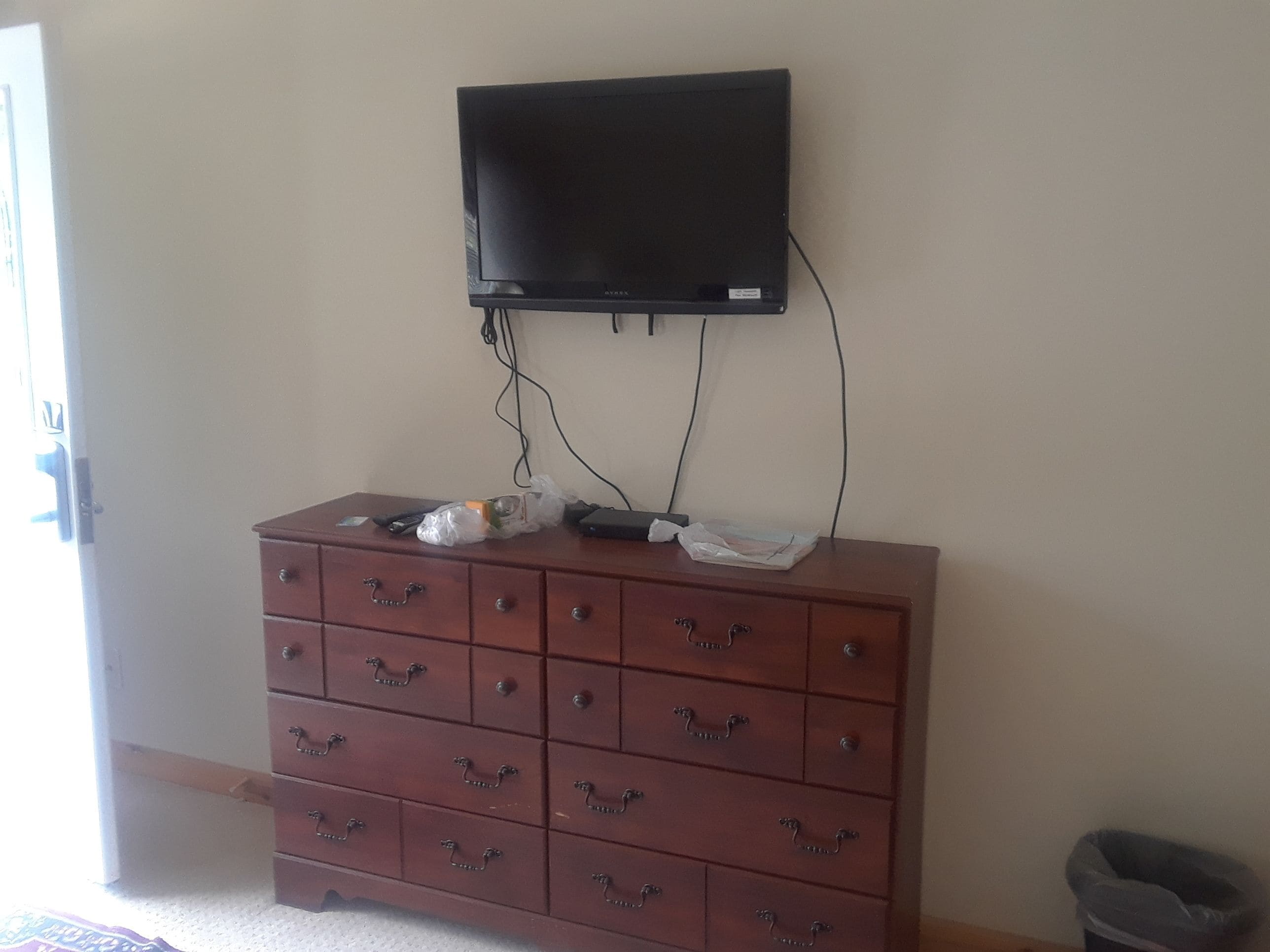 A room with a wooden dresser, a flat-screen TV mounted on the wall, and a trash bin.