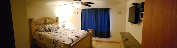 Une chambre à coucher avec un lit, un ventilateur de plafond, un téléviseur fixé au mur et un rideau bleu.