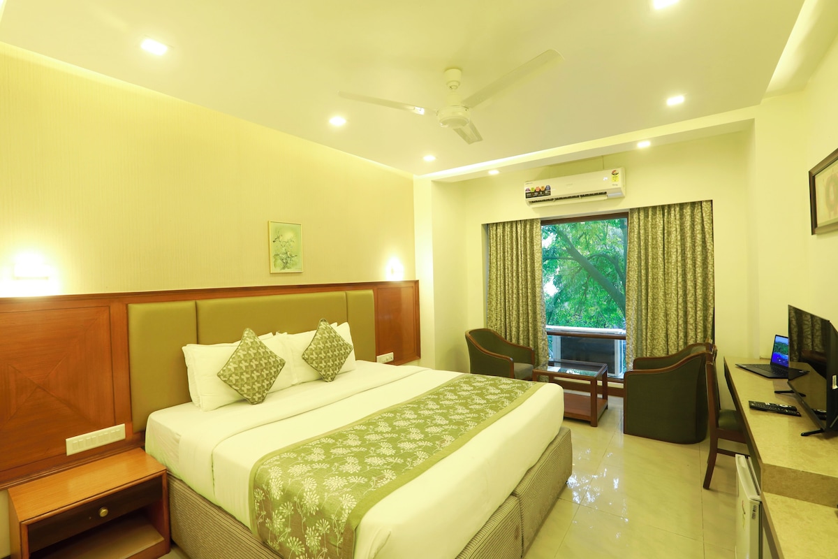 A hotel room with a large bed, a desk with a computer, a chair, and a view of greenery through the window.