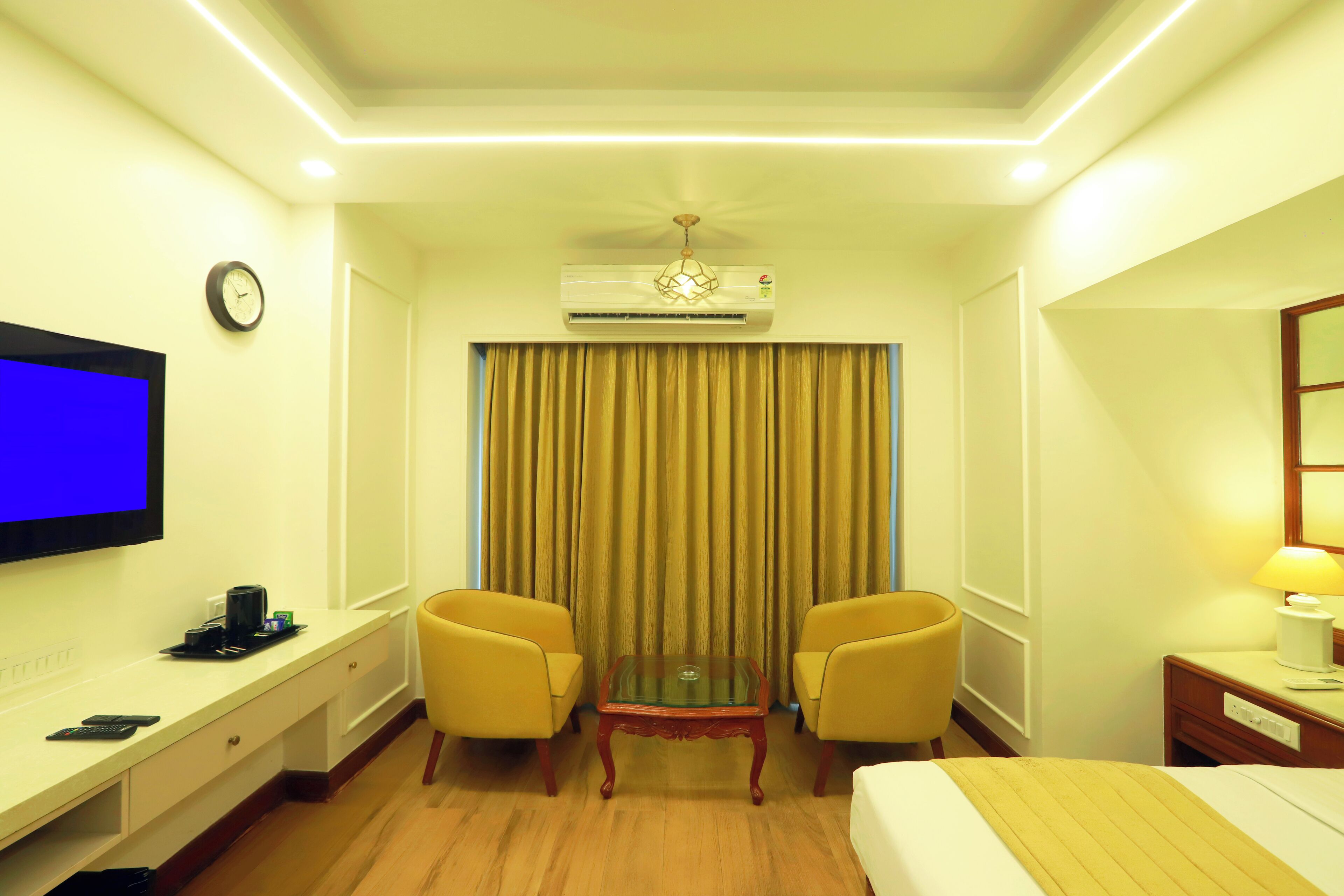 A hotel room with a bed, a desk with a computer, two chairs, a TV, an air conditioner, and a window with curtains.