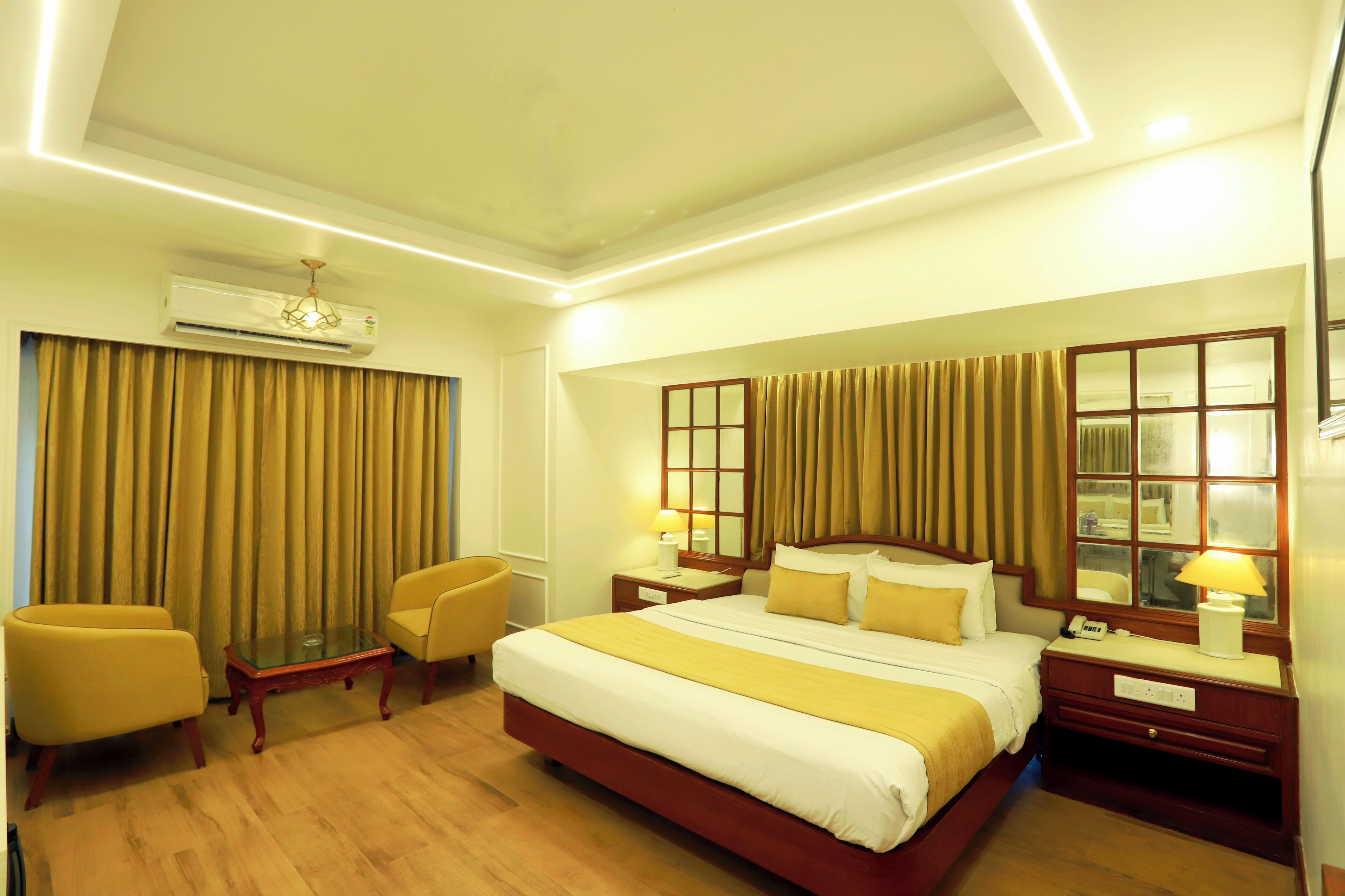 A hotel room with a large bed, two bedside tables with lamps, a small table and two chairs, a mirror, and a window with curtains.