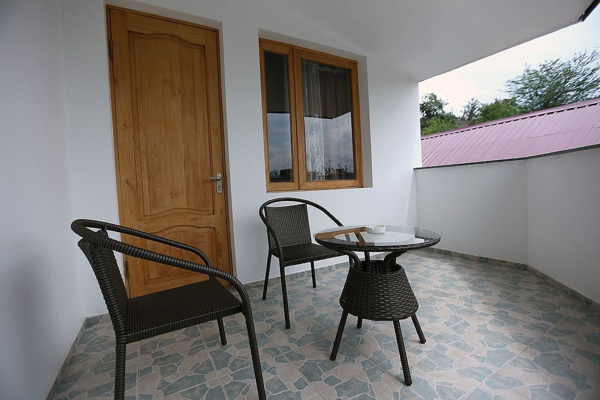 Ein kleiner Balkon mit zwei Korbstühlen und einem runden Tisch, einer Holztür und einem Fenster mit Vorhang.