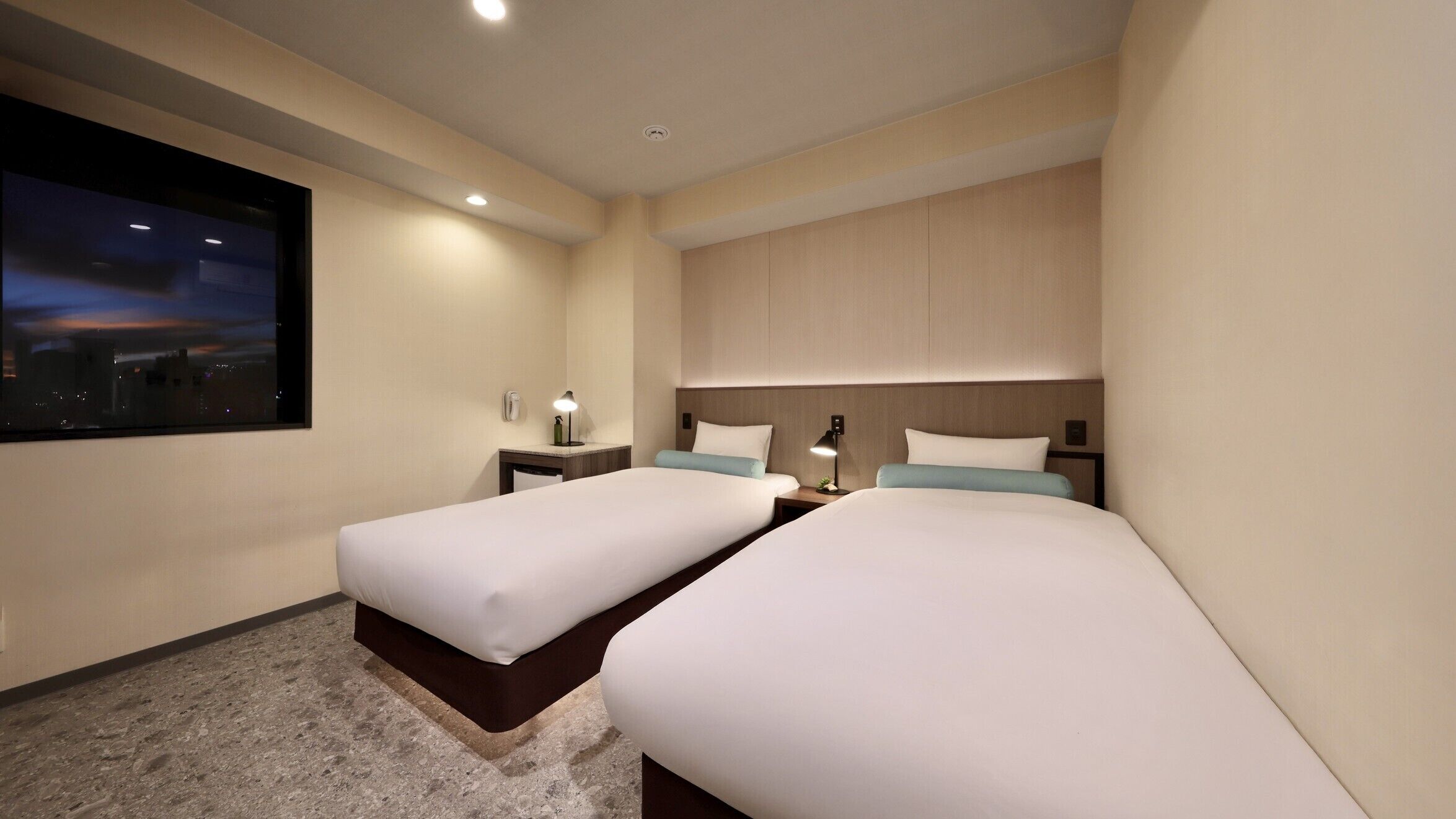 Une chambre d’hôtel avec deux lits, une table de chevet avec une lampe, un téléphone fixé au mur et une grande fenêtre donnant sur le paysage urbain.