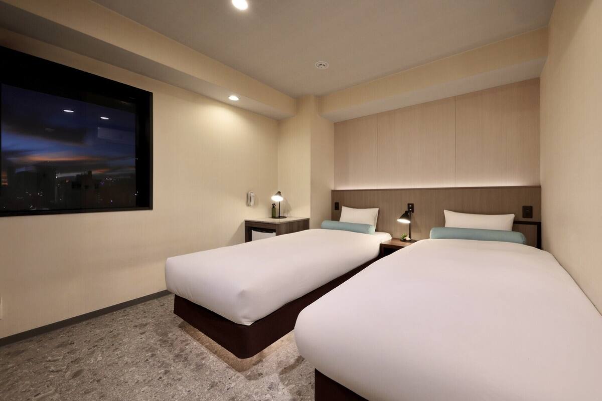 A hotel room with two beds, a nightstand with a lamp, a wall-mounted phone, and a large window with a cityscape view.