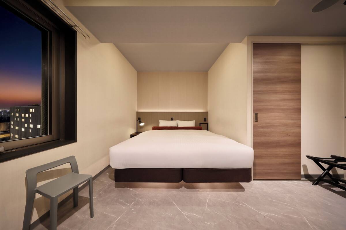 A modern bedroom with a large bed, a nightstand, a chair, and a view of the city at dusk.