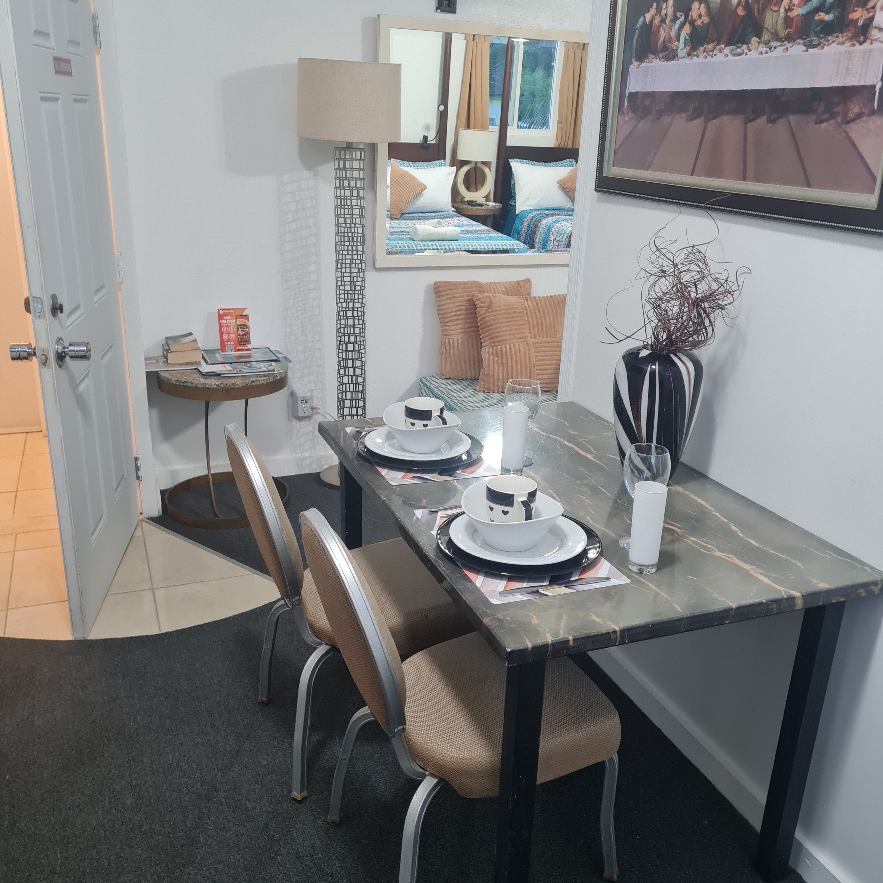 A dining area with a table set for two, a chair, a small round table with a lamp, and a painting on the wall.