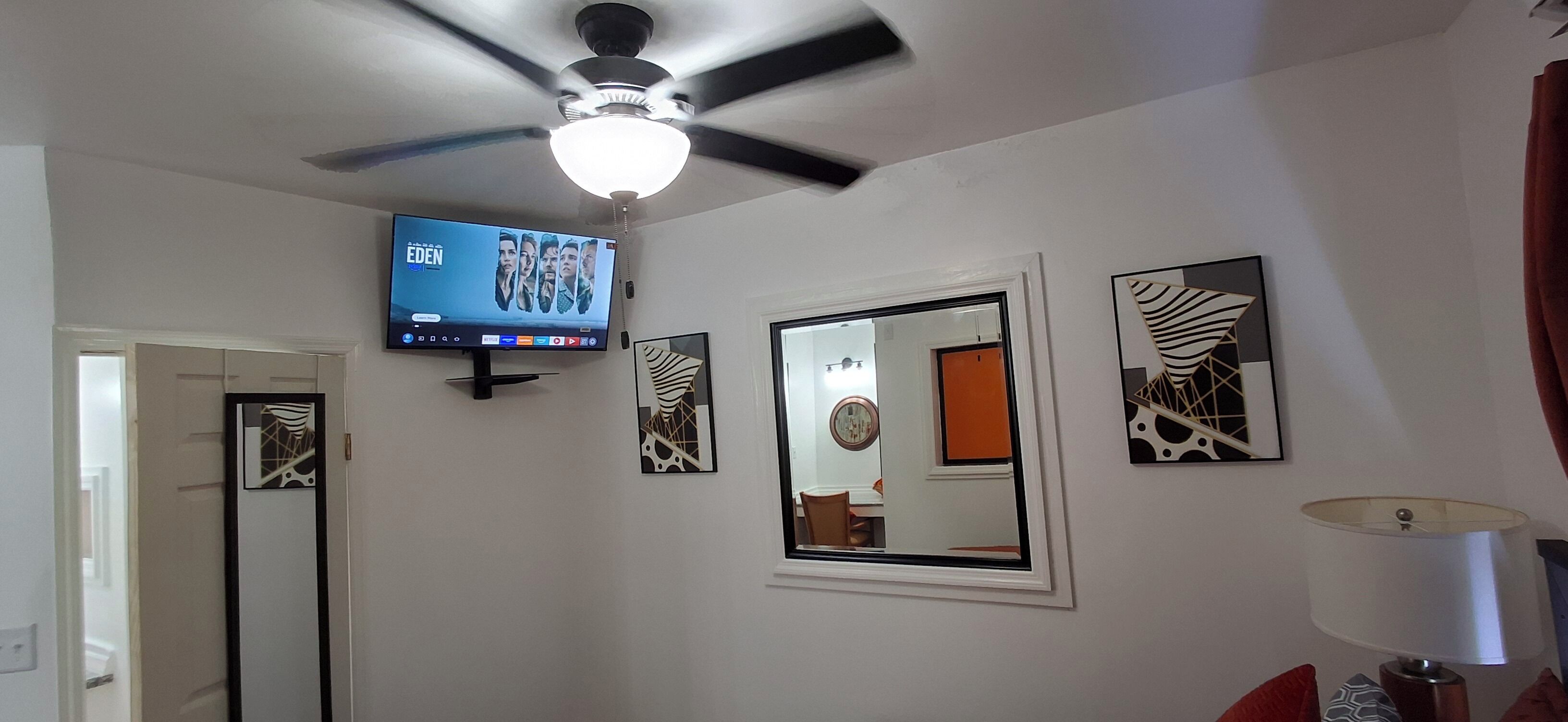 A room with a ceiling fan, a mounted TV displaying 'EDEN', and three wall-mounted mirrors.