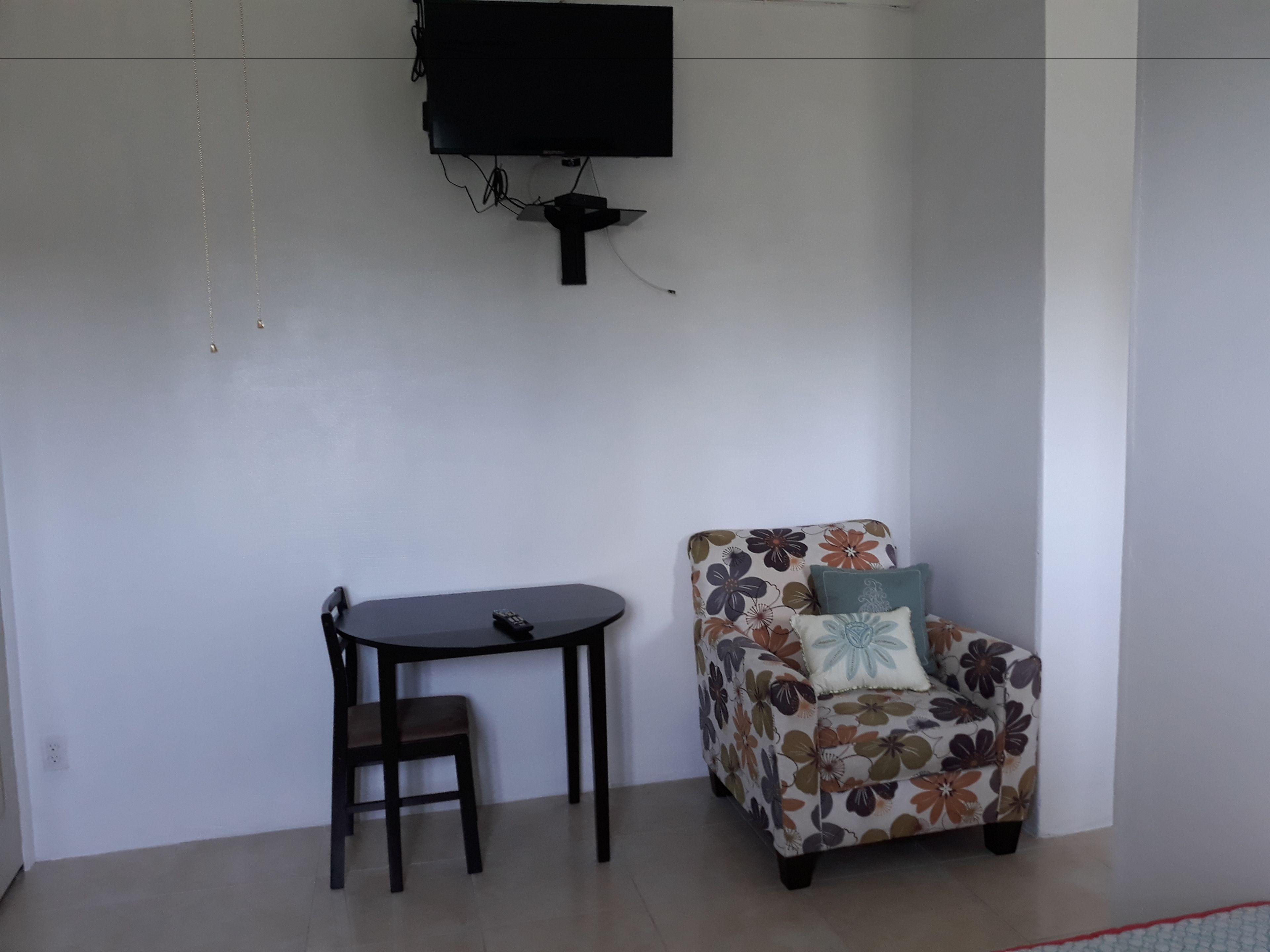 Ein Zimmer mit einem an der Wand montierten Flachbildfernseher, einem kleinen runden Tisch mit Stuhl und einem floralen Sofa mit Kissen.