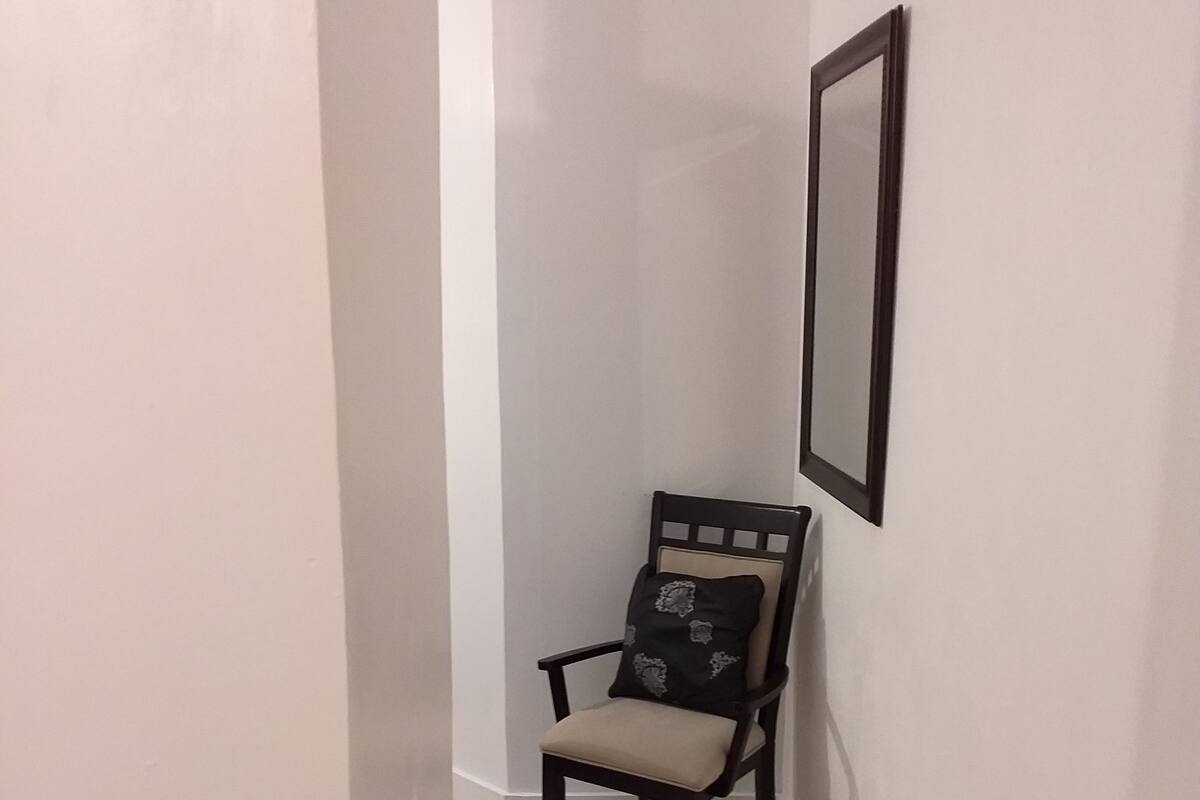 Un pasillo estrecho con una silla de madera oscura con cojín, un espejo en marco en la pared y un suelo de baldosas.