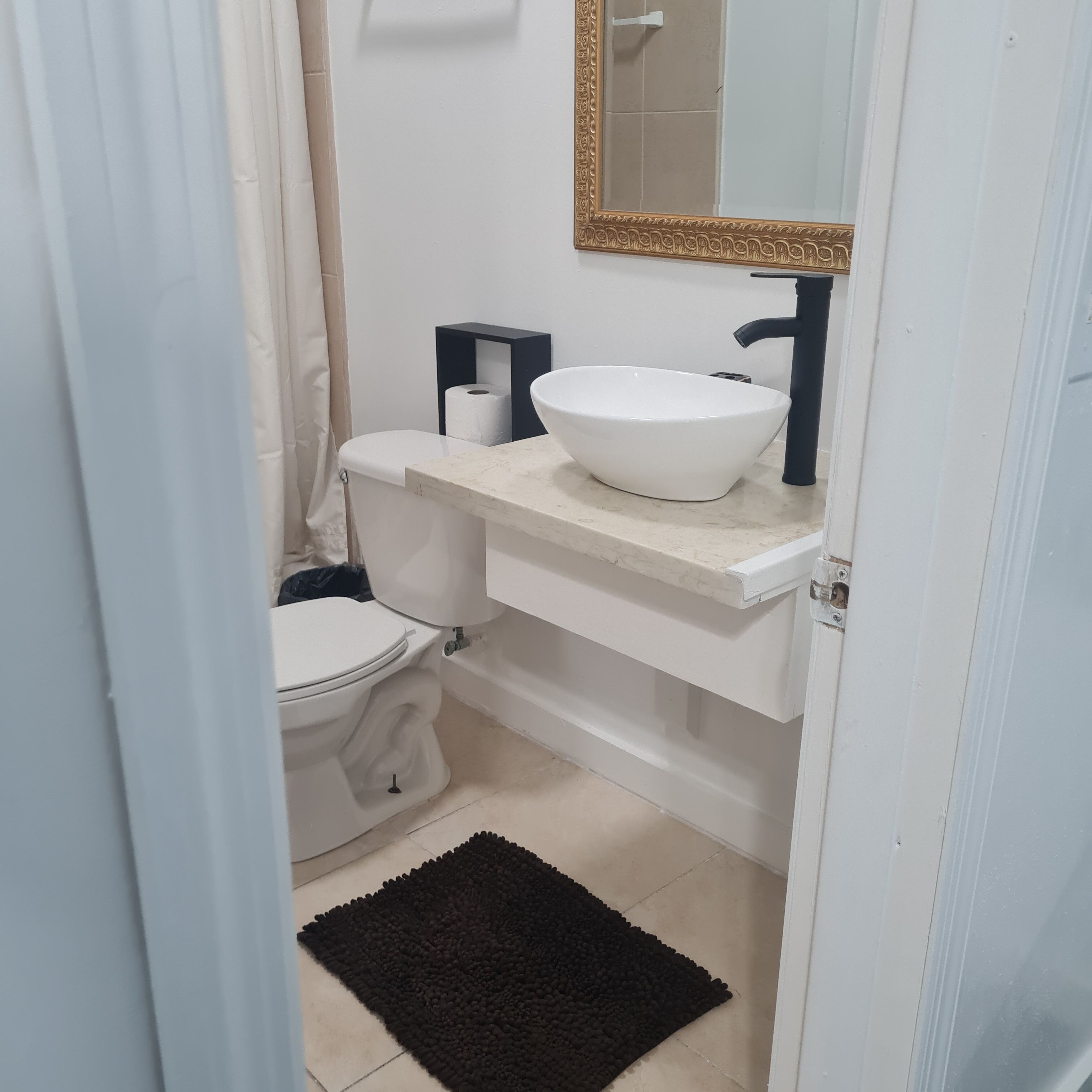 A bathroom with a white toilet, a white sink with a gold-framed mirror above it, and a black rug on the floor.