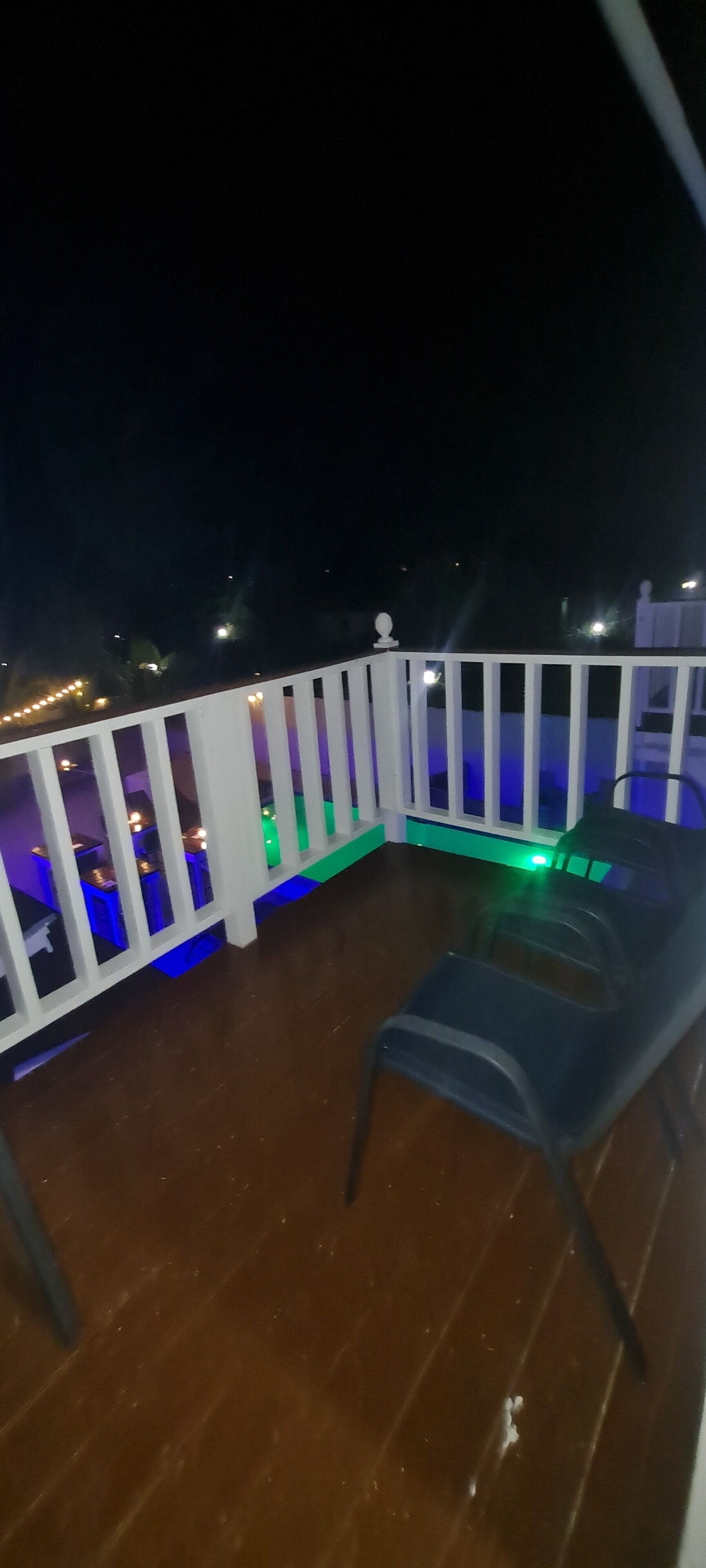 Ein Balkon mit weißer Reling, einem Plastikstuhl und einem Tisch mit einem grünen Licht darunter.