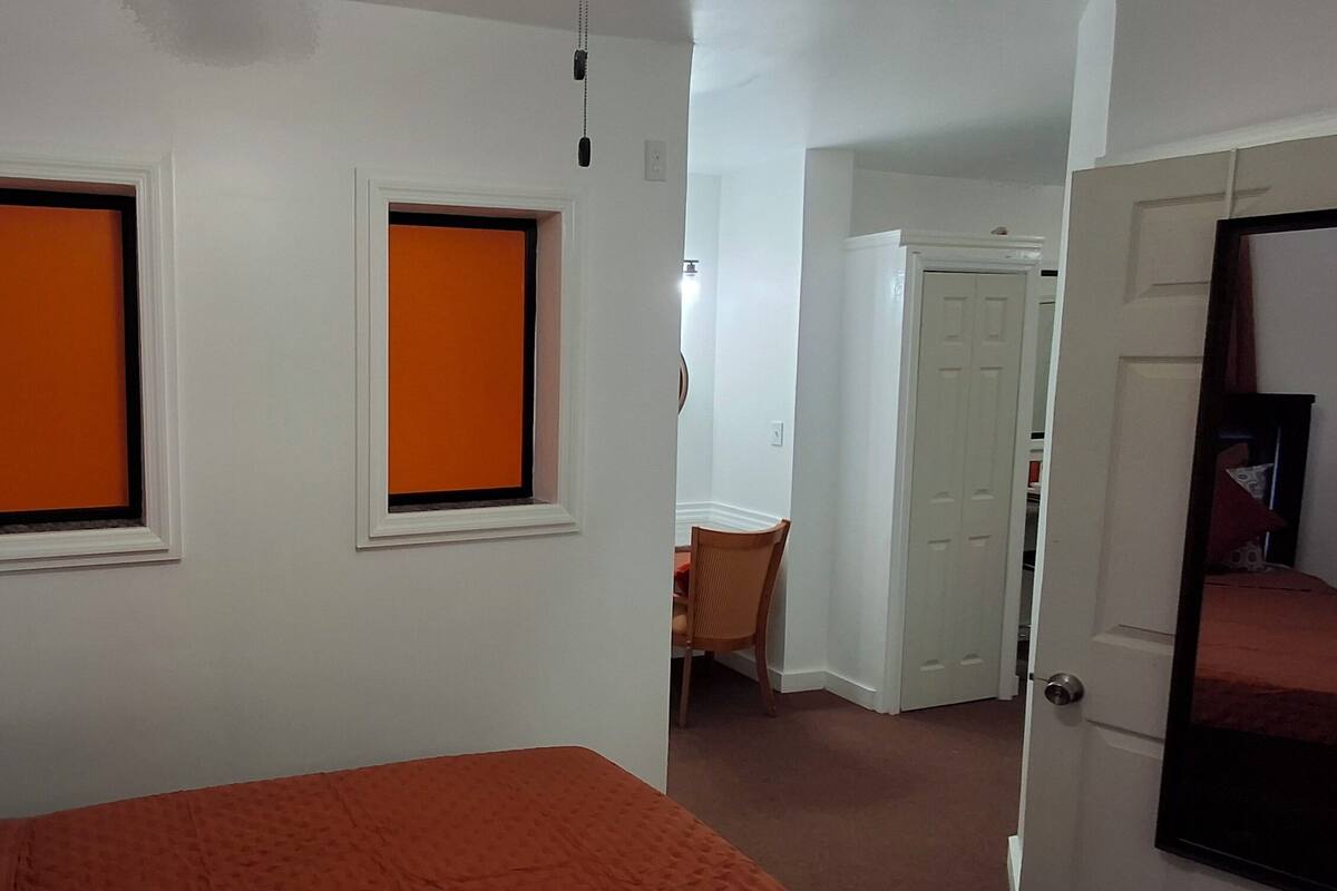 Habitación de hotel con una cama, dos ventanas con persianas naranjas, una silla y una puerta que da a otra habitación.