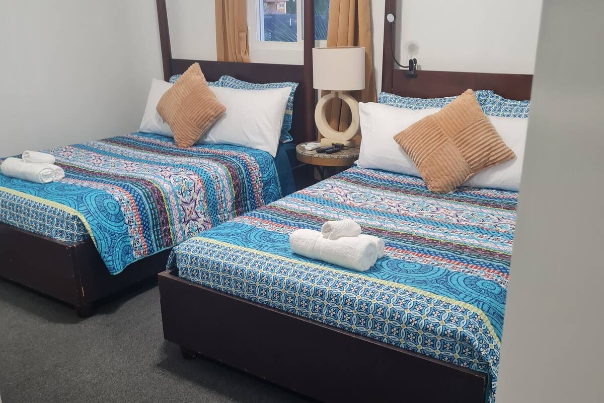 Dos camas individuales con ropa de cama estampada, una mesita de noche con lámpara, y vistas al exterior a través de una ventana.
