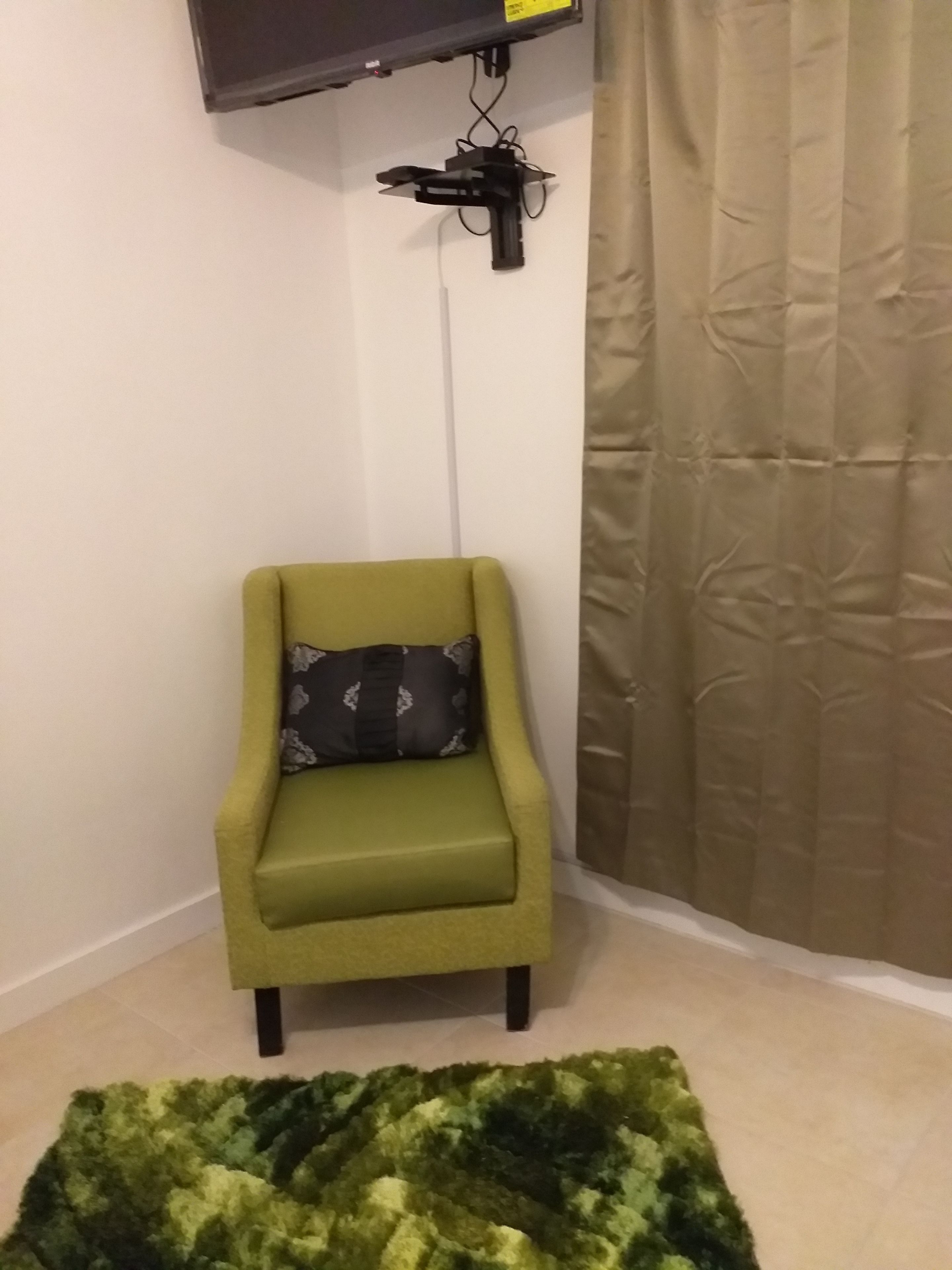A small green armchair with a patterned pillow, a flat-screen TV mounted on the wall, and a beige curtain.
