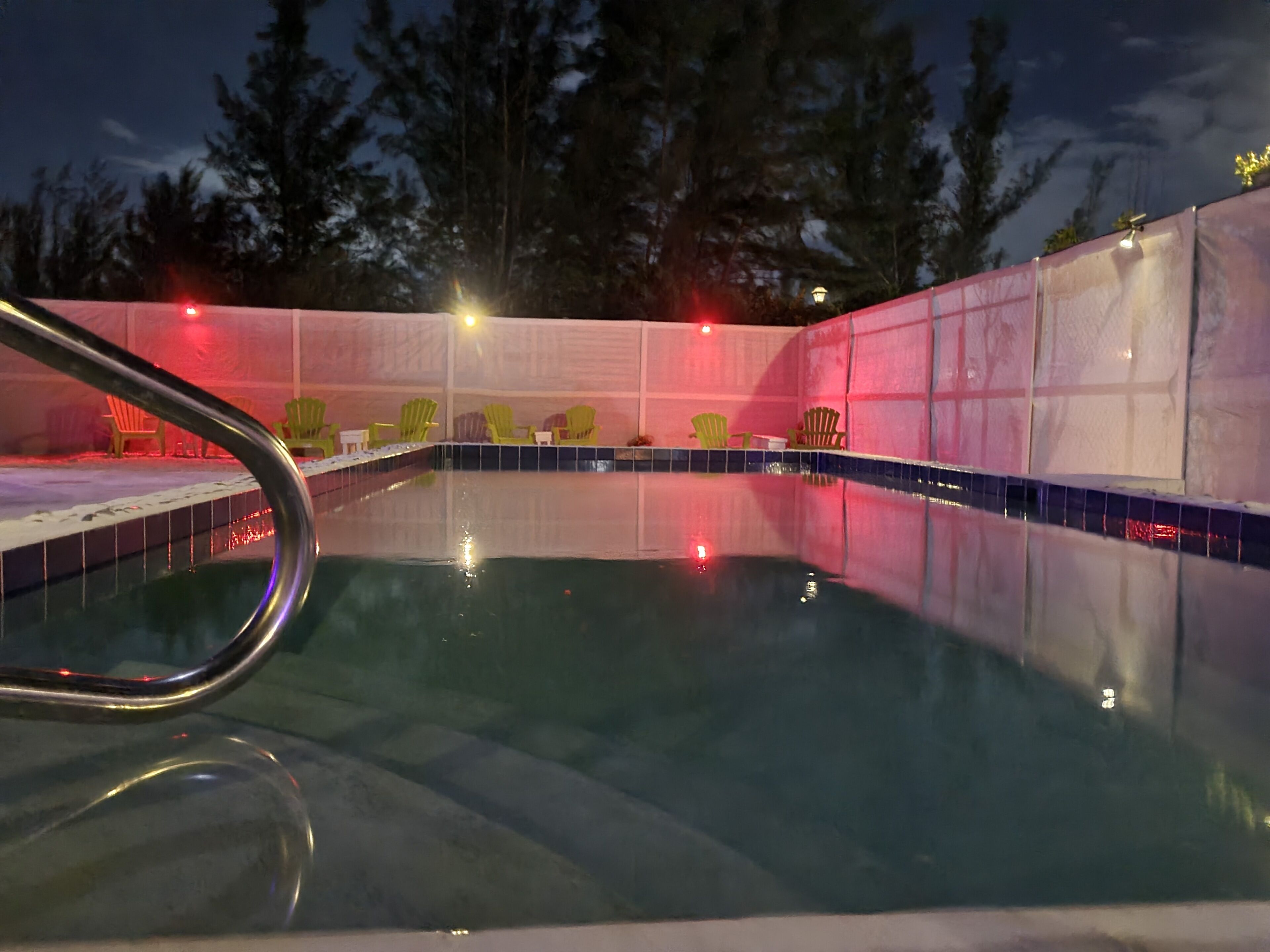 A swimming pool with a metal handrail, surrounded by a pink wall and green chairs.