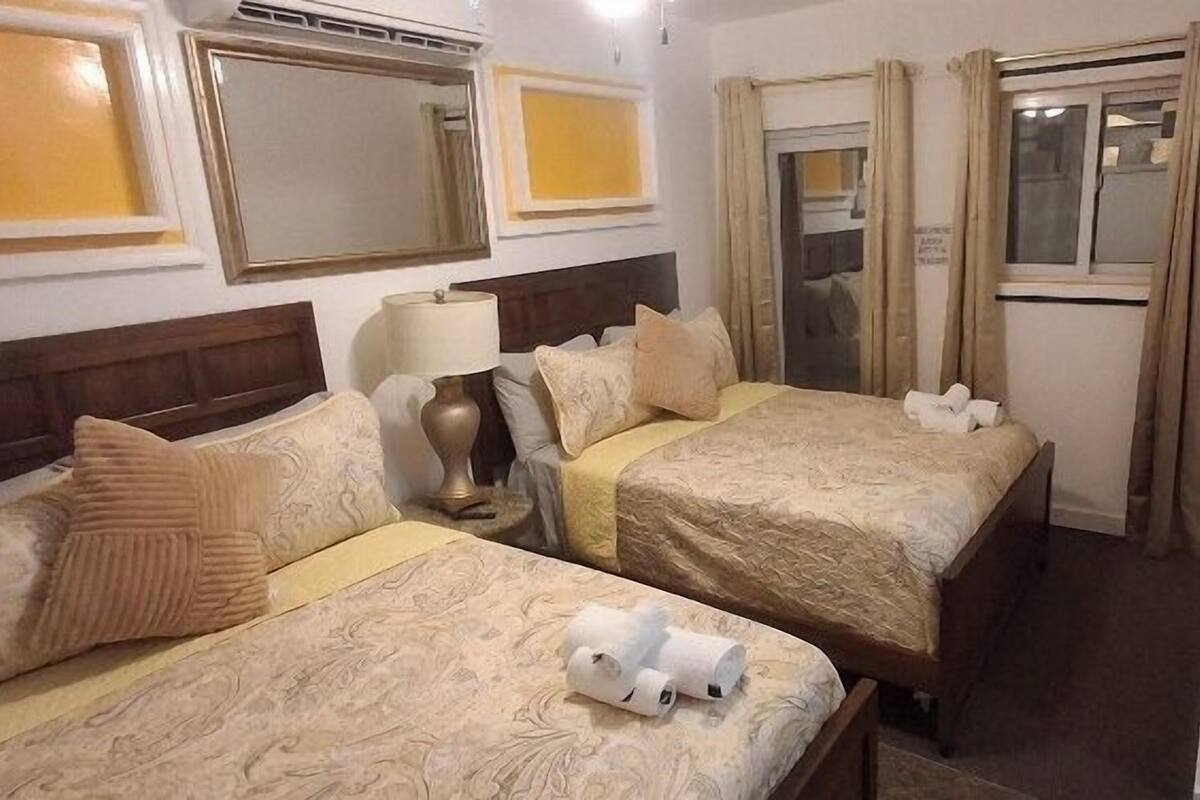 Una habitación de hotel con dos camas, una mesita de noche, una lámpara y un ventanal con cortinas.