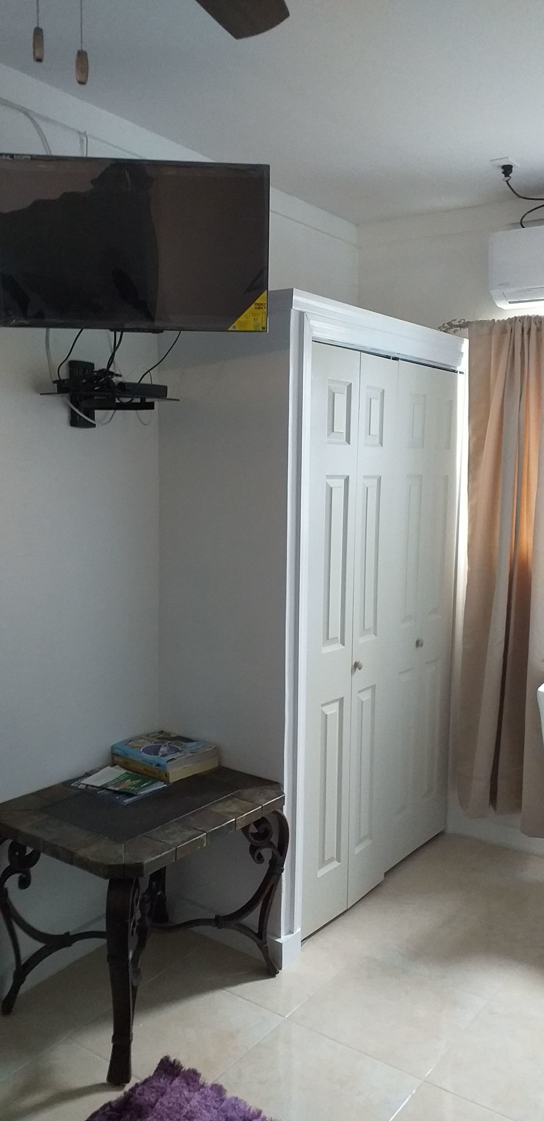 Ein Zimmer mit einem an der Wand befestigten Fernseher, einem weißen Kleiderschrank, einem kleinen Tisch mit Büchern und einem violetten Teppich auf dem Boden.