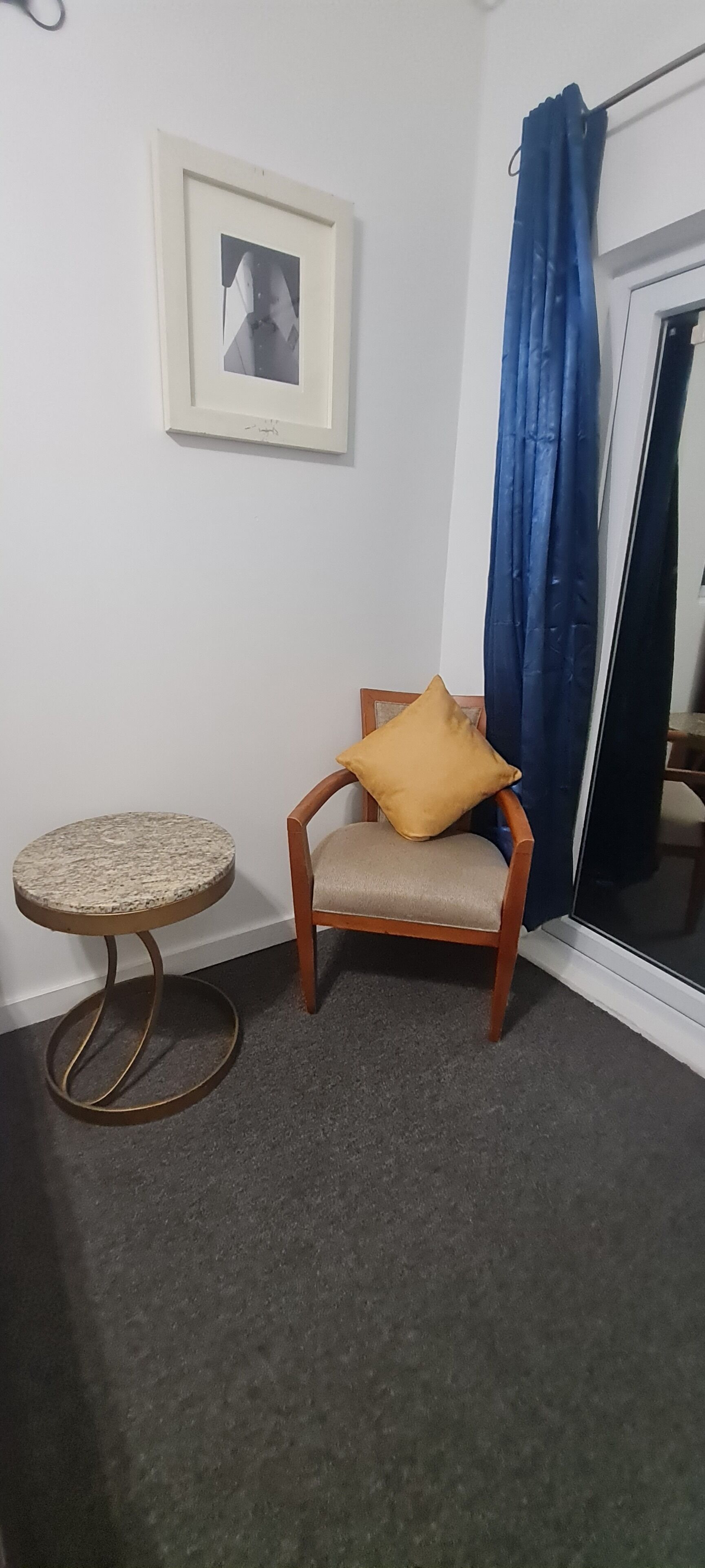 Una habitación con una silla de madera, una mesita redonda pequeña, un cuadro en la pared y una cortina azul.