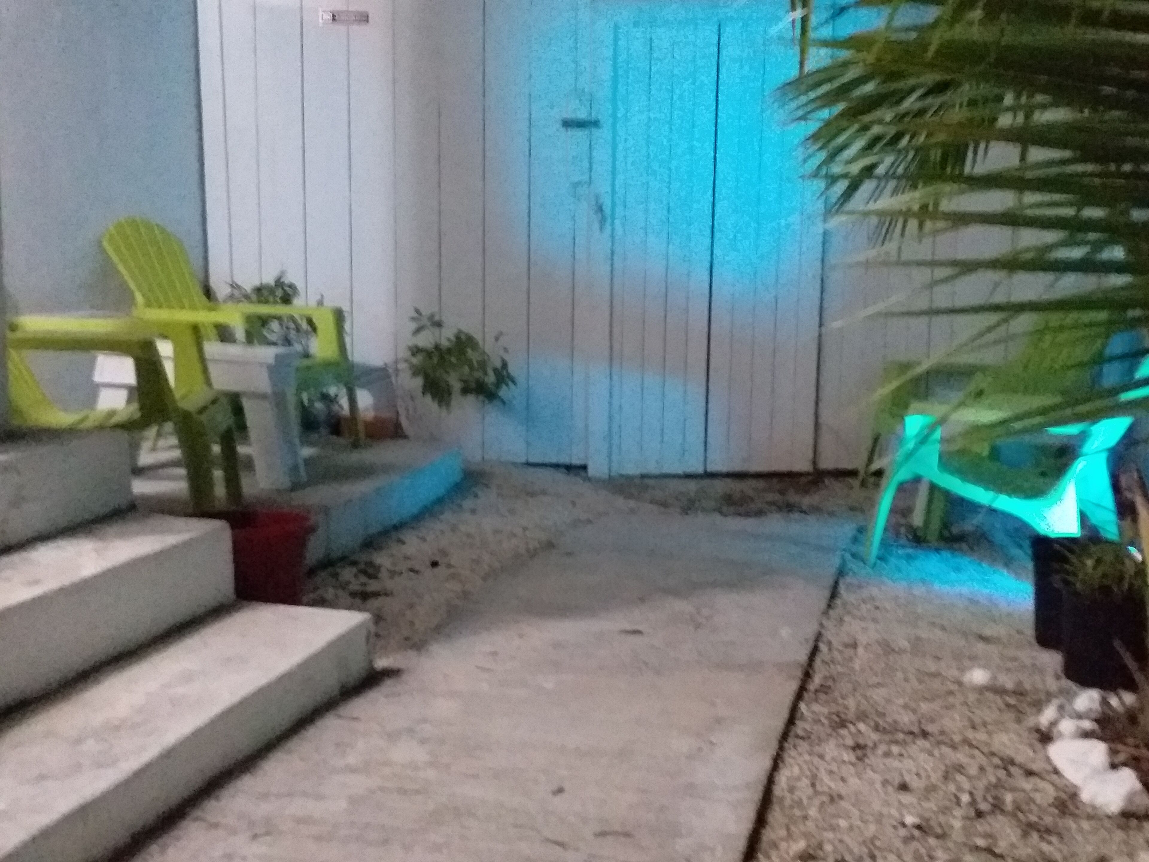 Un sendero de concreto lleva a una puerta blanca con un brillo azul. Hay dos sillas de plástico verde y una planta en maceta cerca de la puerta.
