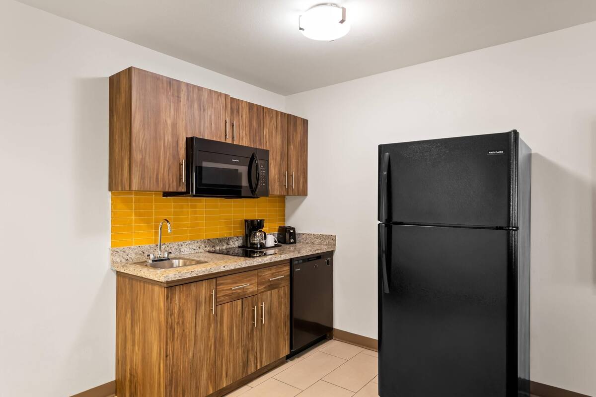 A compact kitchen with wooden cabinets, a black refrigerator, a microwave, and a sink with a yellow backsplash.