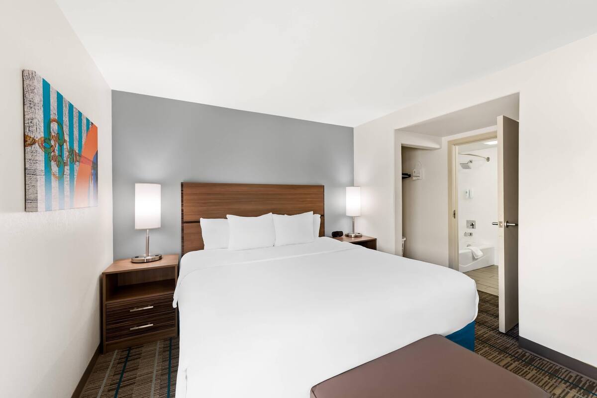 A hotel room with a large bed, two bedside tables with lamps, a wooden headboard, a small bench, a bathroom visible through an open door, and a colorful abstract painting on the wall.