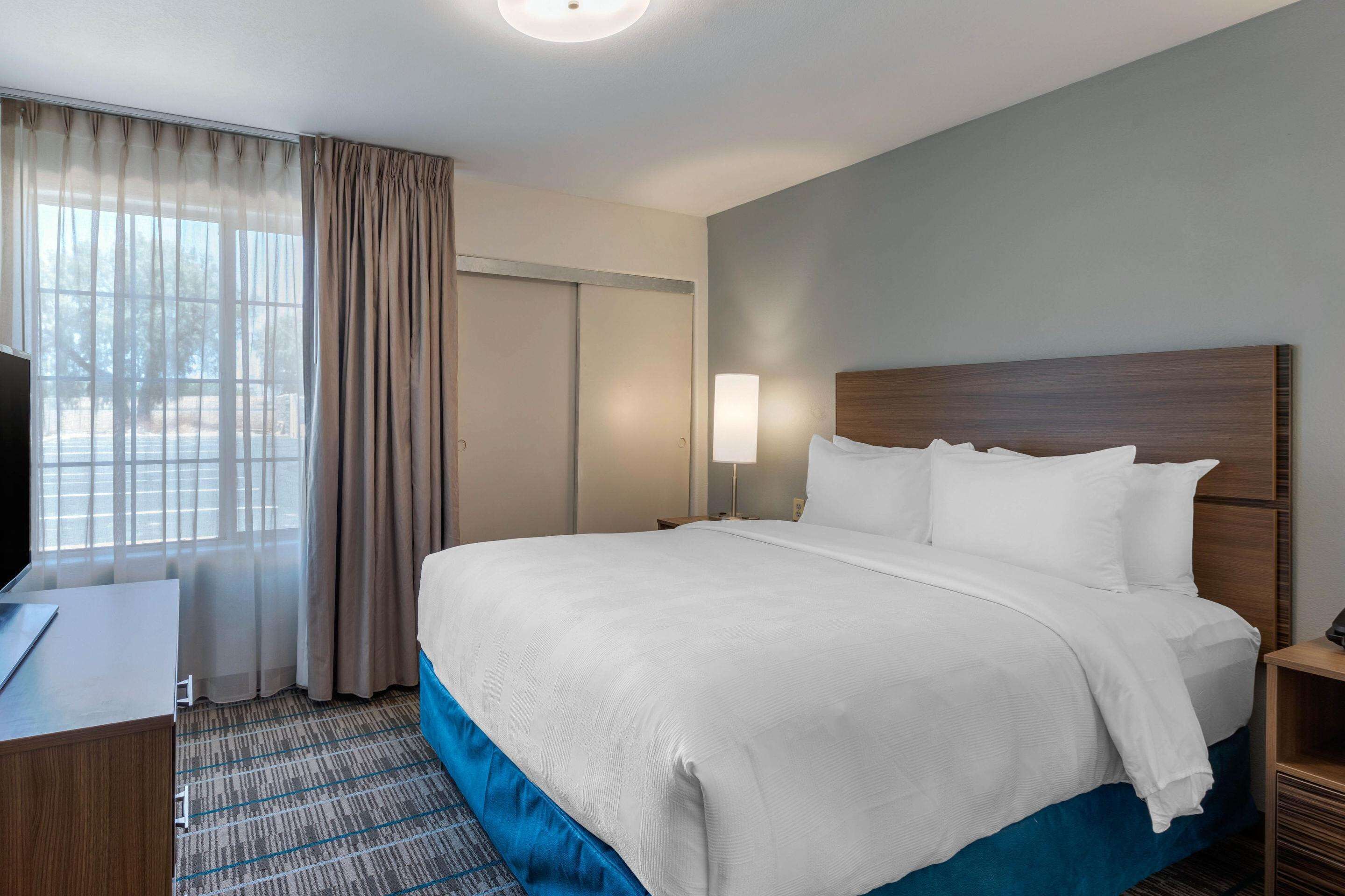 A hotel room with a large bed, a desk, a lamp, and a view of the outdoors through the window.