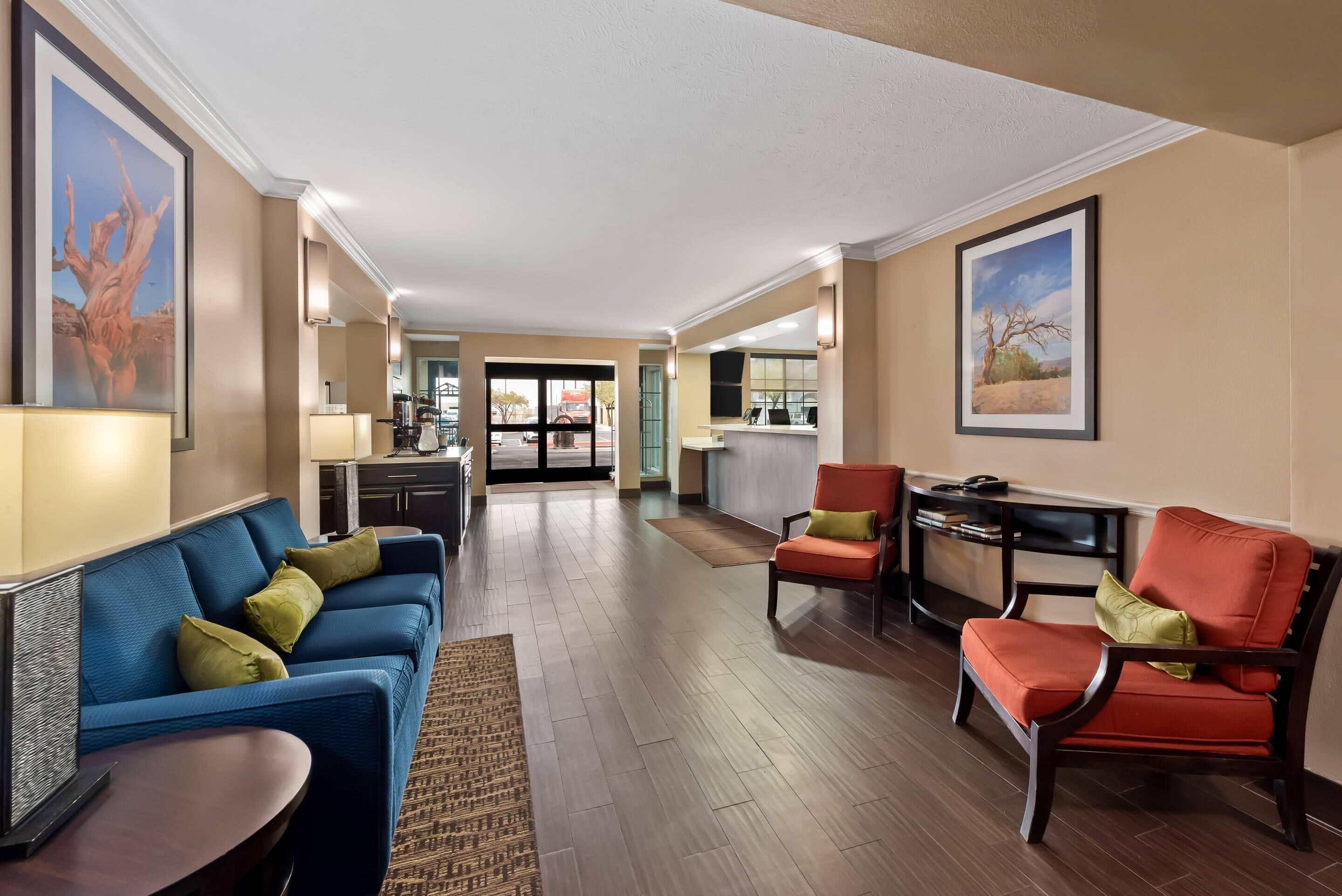A spacious hotel lobby with a blue sofa, red armchair, and a small table. There is a kitchen area in the background.