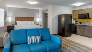 Camera d'albergo con un letto, una poltrona blu, un angolo cottura con frigorifero e microonde, e un comodino con lampada.