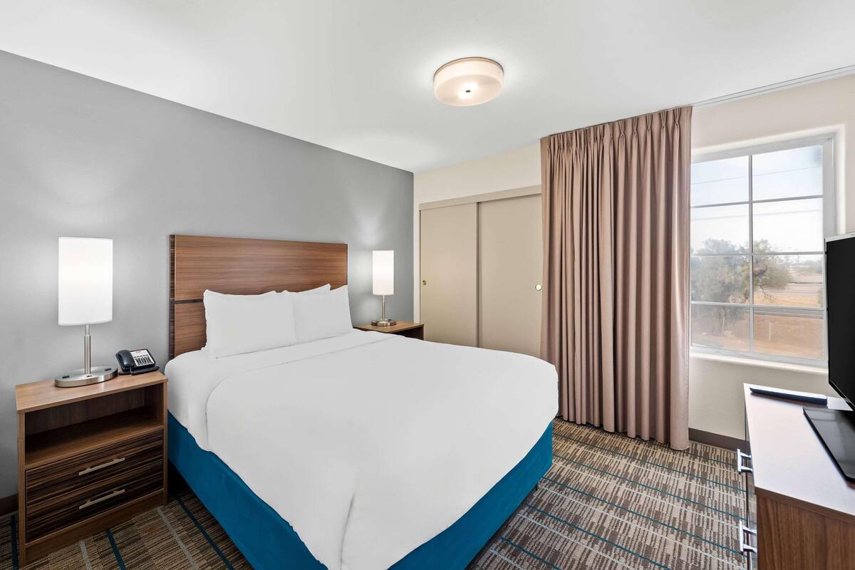 A hotel room with a large bed, bedside tables with lamps, a desk with a computer, and a window with curtains.