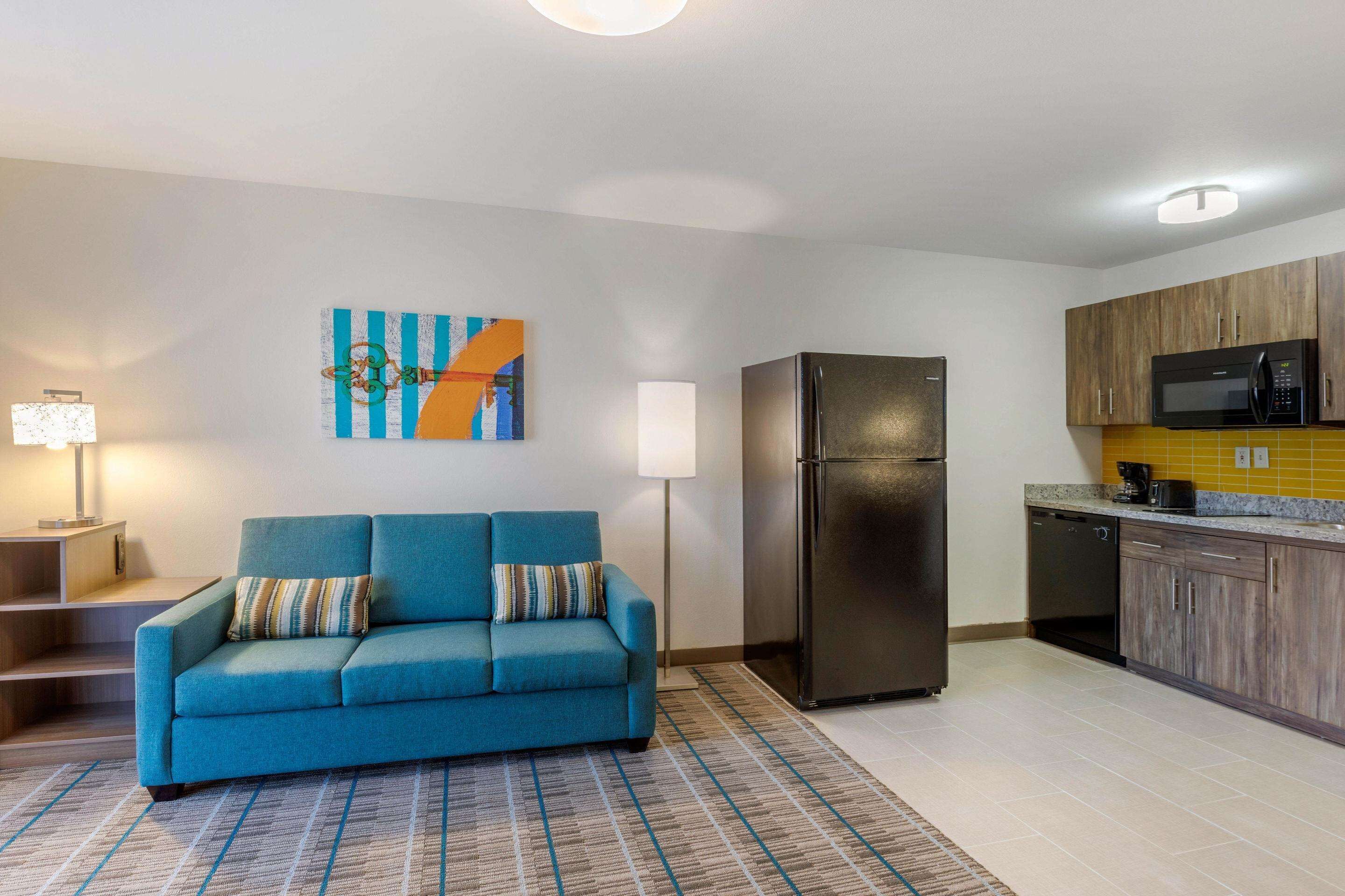 A modern living room with a blue sofa, a refrigerator, a microwave, and a kitchen area with wooden cabinets.