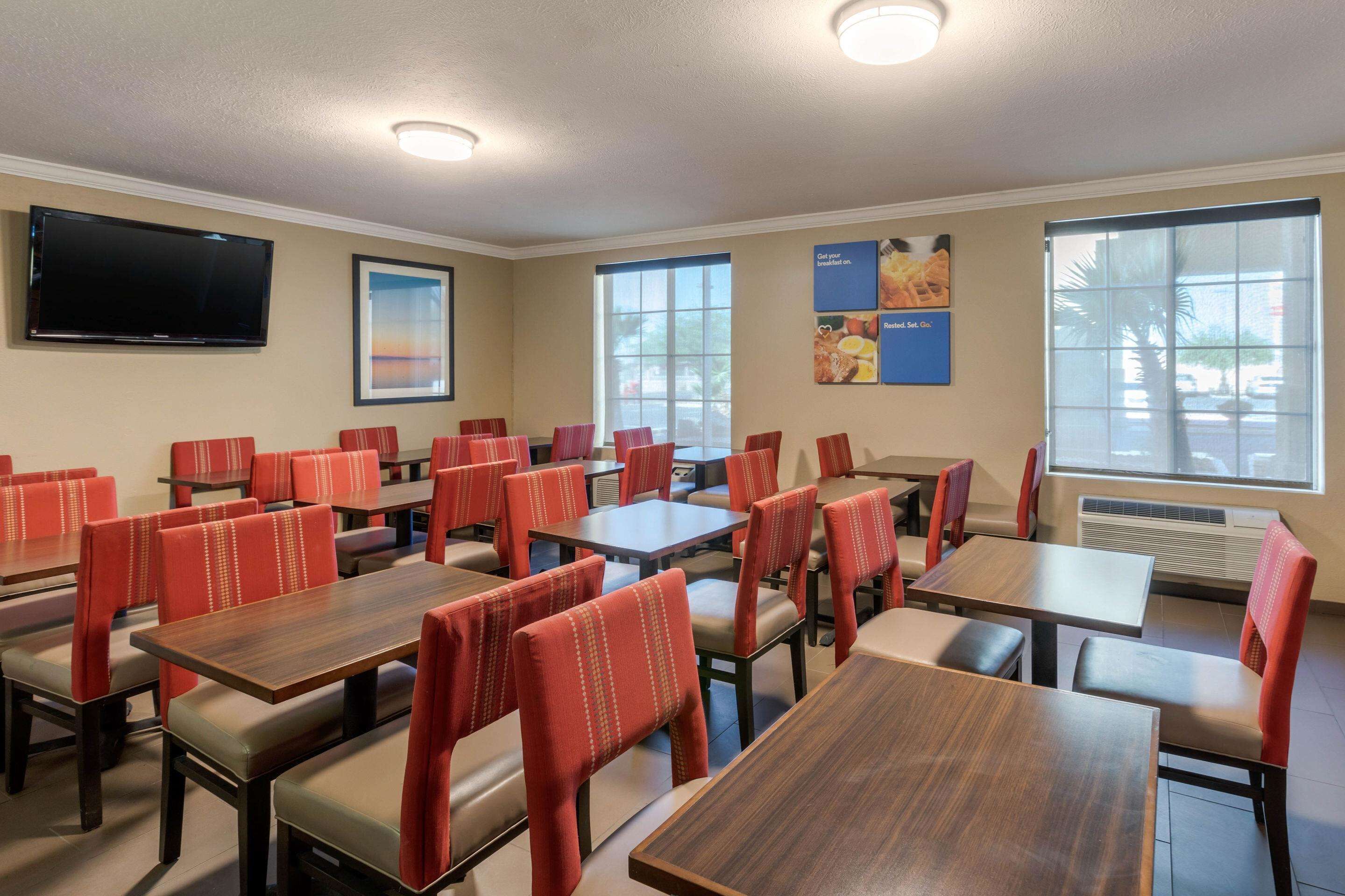 A dining area with multiple tables and chairs, a flat-screen TV mounted on the wall, and large windows providing natural light.