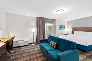 A hotel room with a bed, a blue sofa, a desk with a lamp, and a wall-mounted air conditioner. - Mainstay Suites El Centro I-8 (El Centro)