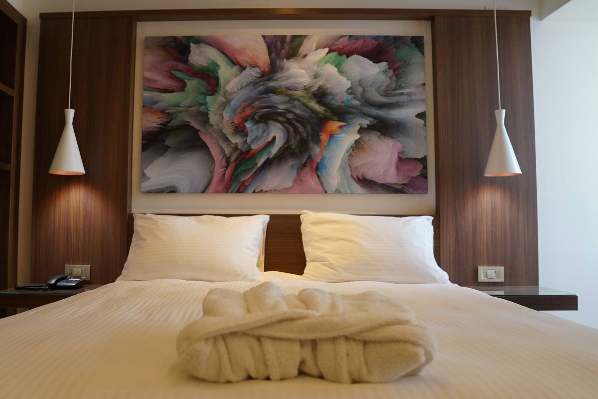 A hotel room with a large bed, two bedside lamps, a painting above the bed, and a wooden wardrobe.