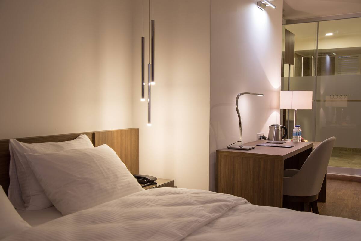 A hotel room with a bed, a desk with a lamp, a chair, and a view of the outside.