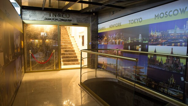 A hotel entrance with a staircase leading to the first floor, a glass railing, and a wall displaying cityscapes of Istanbul, New York, Tokyo, and Moscow. - TN & CO Hotel (Mugla)