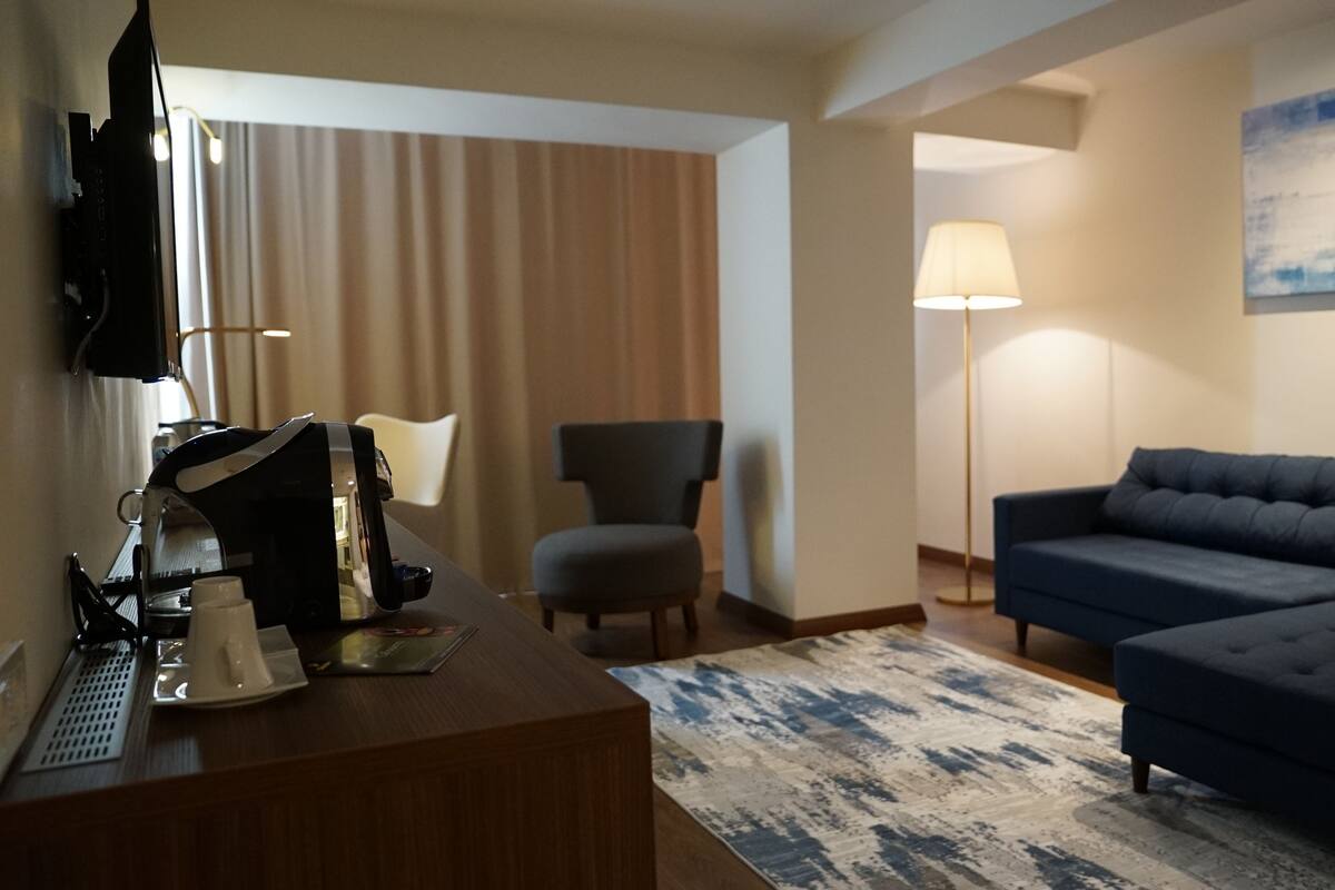 A hotel room with a wooden desk, a sofa, a chair, a lamp, and a painting on the wall.