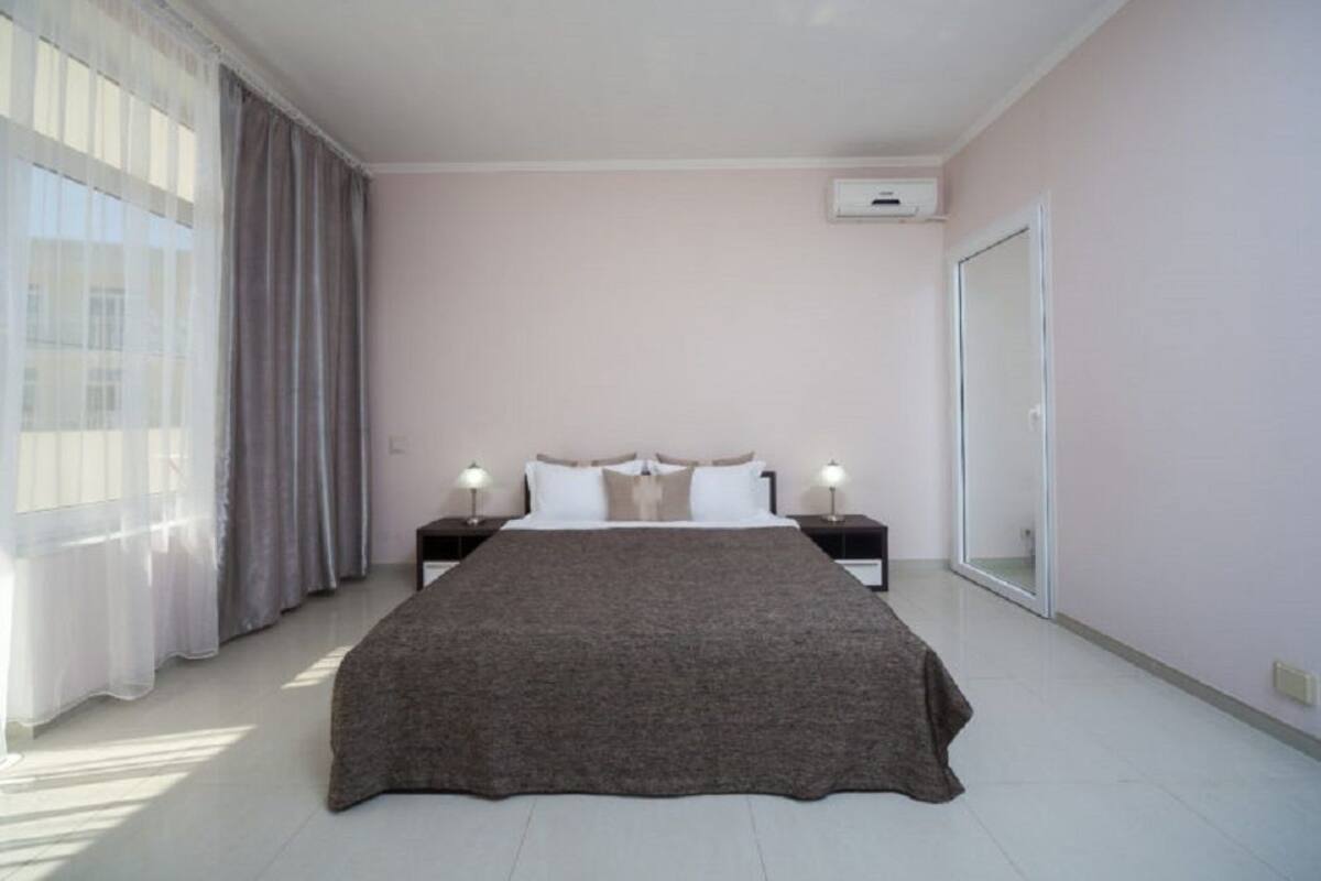 A bedroom with a large bed, two bedside tables with lamps, a window with curtains, and an air conditioning unit on the wall.