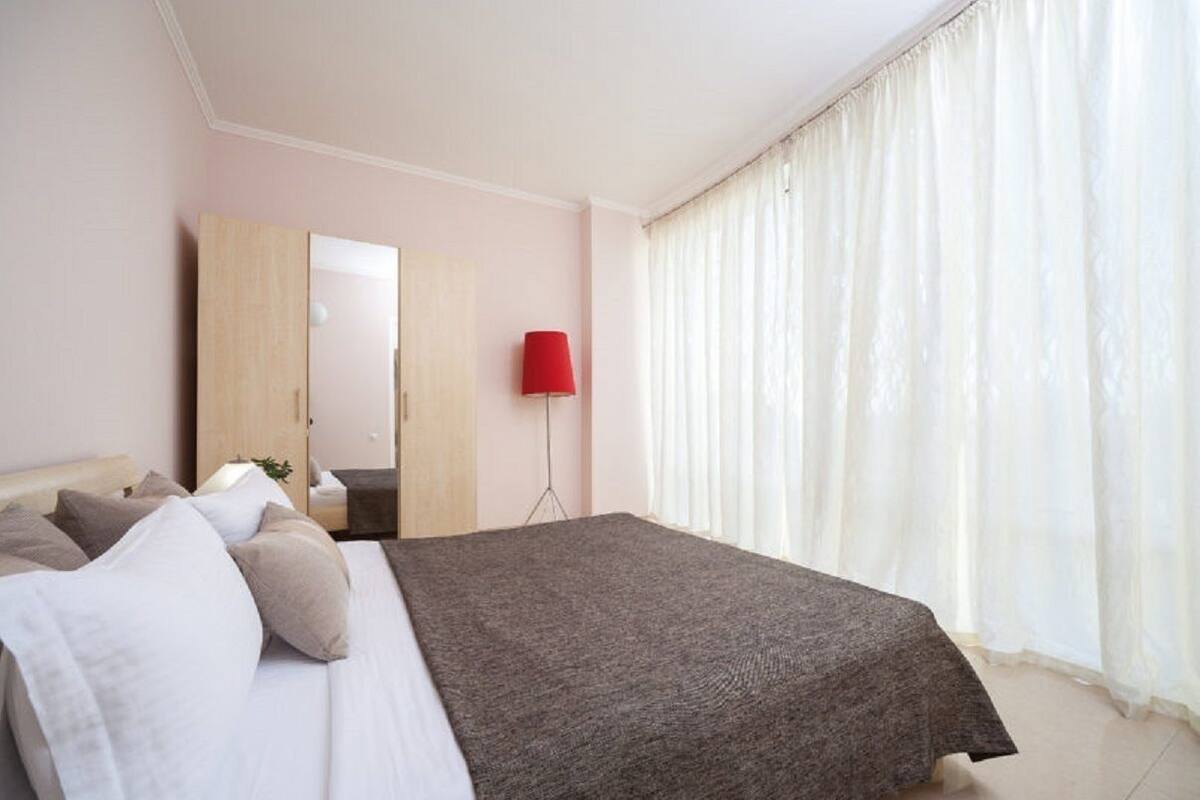 A bedroom with a bed, a mirror, a red lamp, and a window with sheer curtains.