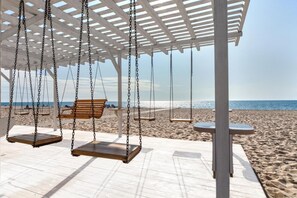 A beachside swing area with wooden swings and a bench, a covered seating area, and a view of the beach and ocean. - Priboy Beach Hotel (Zatoka)