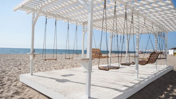 Une balançoire de plage avec des poteaux et des chaînes en bois peints en blanc, installée sur une plage de sable, avec l’océan en arrière-plan.
