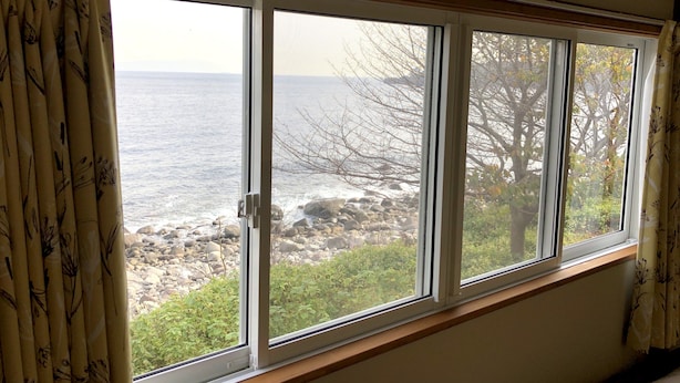 A room with a large window overlooking a rocky shoreline with trees and the sea. - Condominium Seafront Moon and Sand (Ito)