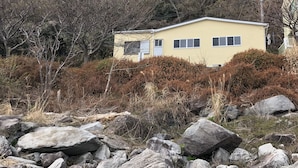 A yellow building surrounded by rocks and bushes, with a forested hill in the background. - Condominium Seafront Moon and Sand (Ito)