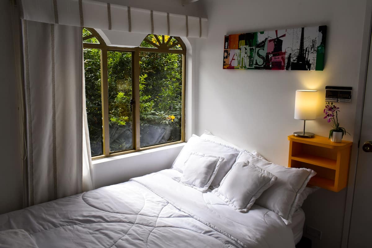 A neatly made bed with white linens, a window with curtains, a wall-mounted shelf with a lamp, and a colorful abstract painting.