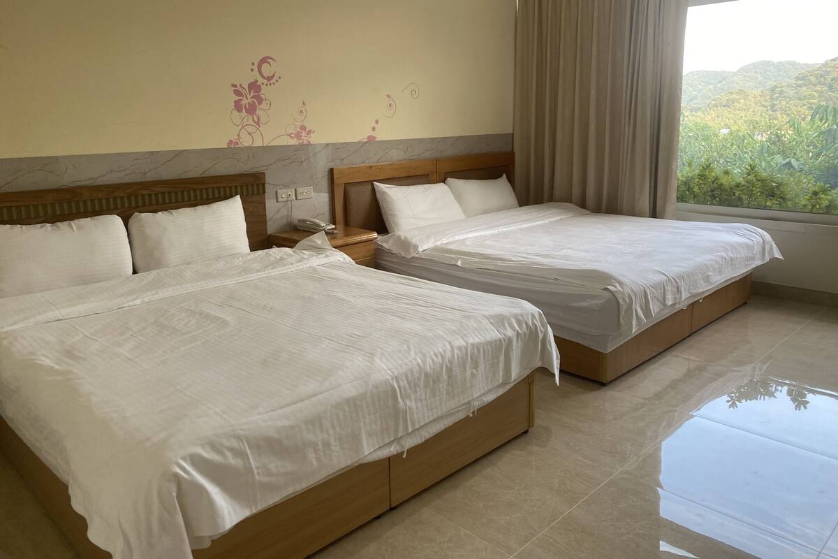 A hotel room with two beds, a nightstand, a telephone, and a window with a view of greenery.