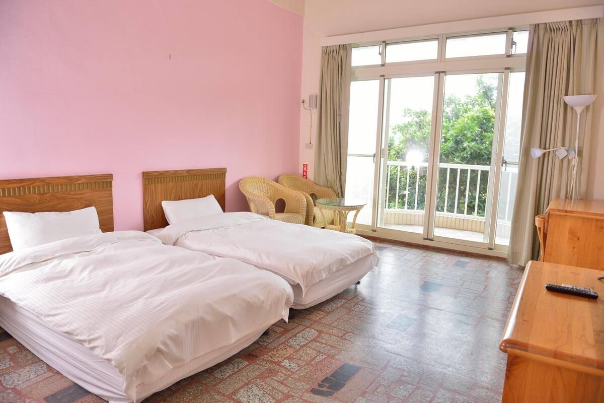 A hotel room with two beds, a balcony with a wicker chair, and a wooden desk.